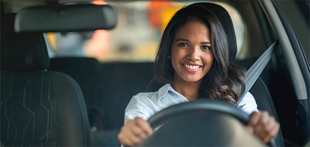 A young woman with long dark hair smiles as she sits in the driver's seat of a car.