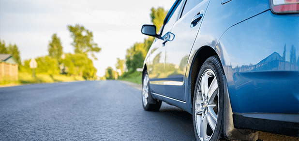 Close-up view of a blue sedan tire driving on a paved road with trees on the sides during a bright afternoon.