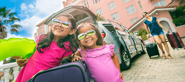 FTwo young girls wearing pink shirts and goggles smile as their mother packs a Dollar car rental in the background.